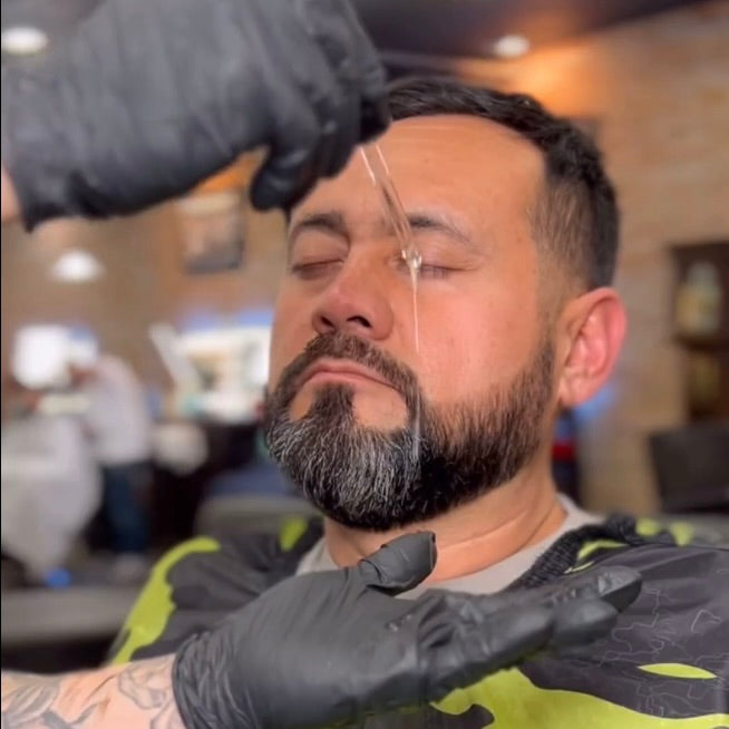 The photo shows a man with a well-groomed beard and closed eyes in a barbershop. A gloved hand is using a dropper to apply El Barber Beard Oil to his beard. The dropper is releasing the oil directly onto his beard, highlighting the grooming process. The setting reinforces the professional use and effectiveness of the beard oil in maintaining a healthy, well-maintained beard.