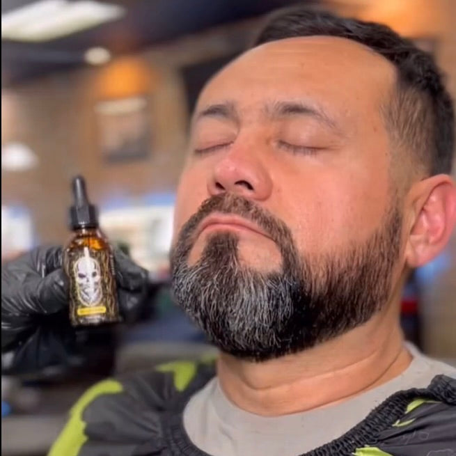 The photo shows a man with a well-groomed beard and closed eyes, sitting in a barbershop. A gloved hand is holding a bottle of El Barber Beard Oil near his face. The bottle is amber with a black dropper cap, and its label features a skull with a beard. The setting suggests that the beard oil is being used or demonstrated as part of a grooming routine.